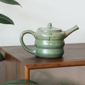 May include: A handmade ceramic teapot with a glossy, mottled sage green glaze. The teapot features a rounded, tiered body, a curved handle on the left side, a straight spout on the right, and a matching lid with a small knob on top.