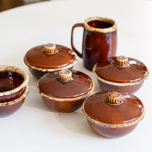 May include: A collection of brown ceramic bowls and a mug. The bowls have lids with a decorative knob. The mug has a handle and a similar brown glaze. The rims have a lighter, speckled finish.