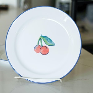 May include: White ceramic plate with a blue rim and a painted cherry design. The cherries are red with green leaves. A second plate is partially visible in the background.