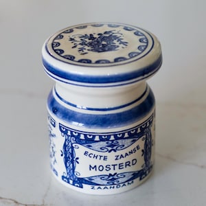 May include: A white and blue ceramic mustard jar with a lid. The jar has a cylindrical shape with a wider base and a narrower neck. The jar is decorated with blue floral patterns and the text "Echte Zaanse Mosterd".
