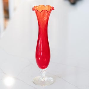May include: A tall, red glass vase with a flared, ruffled orange rim. The vase has a slender, elongated body and sits on a clear, textured base. The vase is likely used for decorative purposes.