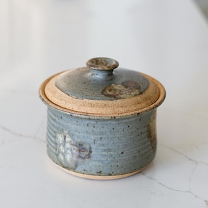 May include: A handmade ceramic lidded jar with a speckled blue-gray glaze and a natural tan rim. The jar has a round shape with a lid and a small knob. The jar is likely used for storing food or other small items.