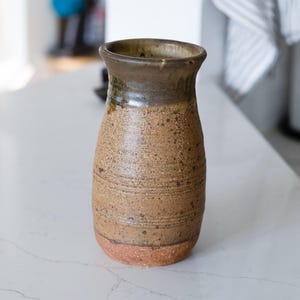 May include: A handcrafted ceramic vase with a textured, speckled surface. The vase features a brown and tan color scheme, with a darker brown rim and a reddish-brown base. The vase is approximately 7 inches tall.