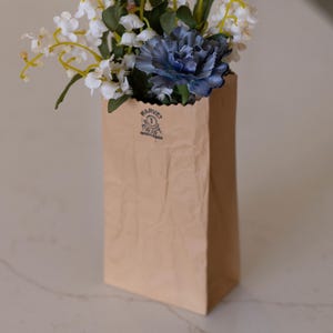 May include: A brown paper bag vase filled with white and blue flowers. The bag has a logo printed on it. The flowers are a mix of white, yellow, and blue, creating a natural and elegant arrangement. The vase is sitting on a white surface.