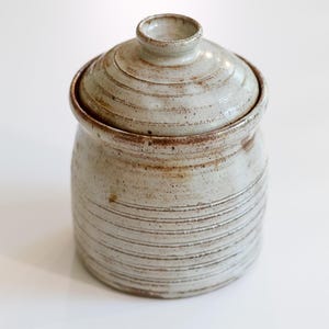 May include: A handmade ceramic jar with a lid. The jar is a light beige color with horizontal textured lines and a brown rim. The lid has a small knob on top. The jar is likely used for storage.