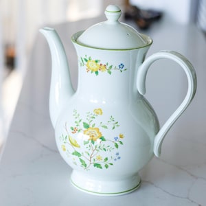 May include: White ceramic teapot with a lid and a curved handle. The teapot is decorated with yellow and blue floral patterns and green trim. The teapot is sitting on a white surface.