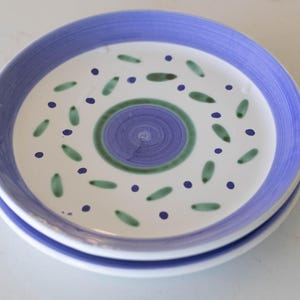 May include: Two stacked ceramic plates with a white center and a blue rim. The plates are decorated with green leaf shapes, blue dots, and a central spiral design in blue and green. The plates are suitable for serving food.