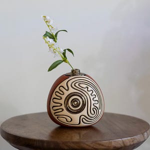 May include: A decorative, egg-shaped vase with a brown and cream color scheme. The vase features a unique, swirling design in black. A small bouquet of white flowers with green leaves is placed inside the vase. The vase is sitting on a wooden table.