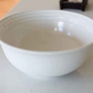 May include: A white ceramic mug with a black handle and the text "But First, Coffee" in a bold, black font. The mug is filled with a dark liquid, likely coffee, and sits on a wooden surface.
