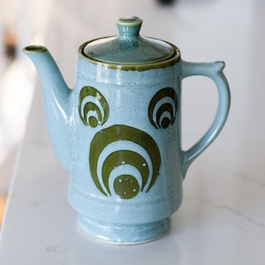 May include: A light blue ceramic teapot with a lid and handle. The teapot has a spout and is decorated with green, concentric, semi-circular designs. The teapot is on a white surface.