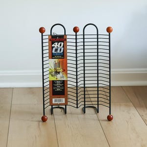 May include: A black metal wire CD storage tower with a capacity for 40 discs. The unit features a vertical design with rounded top arches and small wooden spheres at each of the four corners. A product label is attached, reading Atlantic 40 CD Tower.