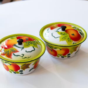 May include: Two white enamel bowls with lids, featuring a vibrant fruit and leaf design in red, orange, and green. Each bowl has a black knob on the lid and a bright green rim. The bowls are on a white surface.