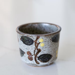 May include: A small, handcrafted ceramic cup with a speckled gray base. The cup features a brown floral design with dark green leaves and yellow accents. The rim is a darker brown, and the cup has a rustic, artisanal aesthetic.