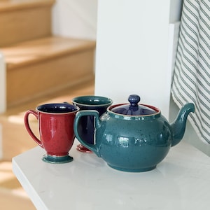 May include: A teal ceramic teapot with a matching lid and a speckled finish, accompanied by two ceramic mugs. One mug is red with a teal base, and the other is dark blue. All pieces feature a smooth, glossy glaze and a sturdy handle design.