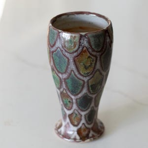May include: A handmade ceramic vase with a unique scale pattern in shades of green, brown, and white. The vase has a flared top and a small, rounded base. The interior is a light beige color.