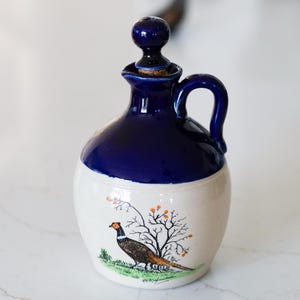 May include: A ceramic jug with a dark blue top and handle, and a cream-colored base. The base features a pheasant illustration with a tree and orange berries. The jug has a dark blue stopper.