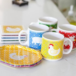 May include: A collection of ceramic mugs and plates with farm animal designs. The mugs feature a white interior and colorful exterior patterns with a cow, pig, and chicken. The plates are yellow with a chicken design. The items are stacked on a white surface.