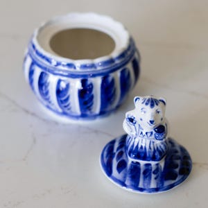 May include: A blue and white ceramic jar with a lid featuring a bear figurine. The jar has a round shape with vertical ribbed detailing. The lid is off, revealing the interior. The bear figurine is on the lid.
