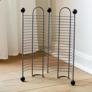 May include: A gray metal magazine rack with a geometric design. The rack has a series of horizontal bars and is supported by rounded feet and topped with dark brown spheres. The rack is positioned on a light wood floor.