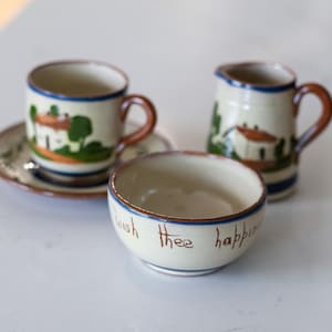 May include: A ceramic tea set featuring a cup and saucer, a creamer, and a small bowl. The set is painted with a landscape design of a house and trees. The bowl has the words "Wish thee happy" written on it.