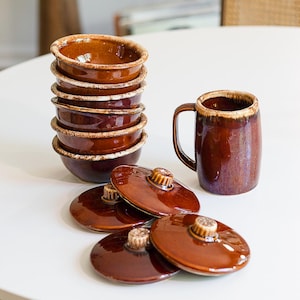 May include: A stack of brown ceramic bowls with lids and a matching mug. The bowls have a rustic, glazed finish with a lighter rim. The lids have a small knob. The mug has a handle and a similar glaze.