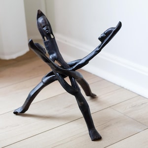 May include: Dark brown wooden sculpture of intertwined figures. The sculpture has a central figure with outstretched arms and two figures on the ends. The sculpture is on a light wood floor.