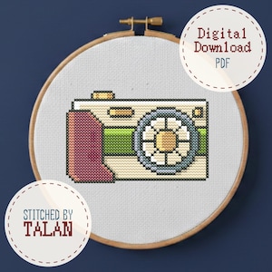 May include: Cross-stitch embroidery of a vintage camera in a wooden hoop. The camera is beige, green, and pink, with a blue and yellow lens. The text "Digital Download PDF" and "Stitched by TALAN" are in the image.