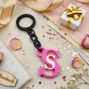 May include: A keychain featuring a black circular ring and a black chain link attached to a bright pink letter S charm. The letter S has a white letter S layered on top of the pink base. The keychain is constructed from plastic components.