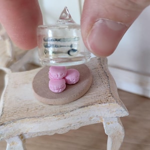 May include: A miniature white table with a glass cloche and pink macarons on a small wooden plate. The cloche has a clear glass lid with a pointed top.