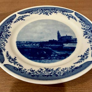Puede incluir: Plato de cerámica blanca con un borde floral azul y blanco y una imagen central de un edificio y un paisaje. El plato tiene un detallado patrón floral azul alrededor del borde y una escena de paisaje azul y blanco.