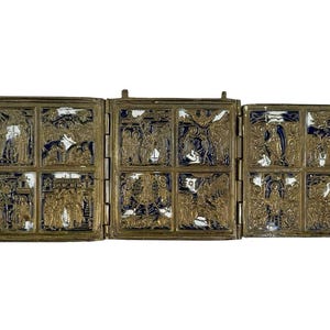 May include: A hinged, gold-toned metal triptych with intricate relief carvings. Each panel is divided into six smaller squares, each depicting a religious scene with figures and architectural details. The background of each square is a dark blue color.