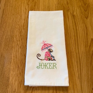 May include: White hand towel with an embroidered monkey wearing a pink and red outfit and holding a pink umbrella. The word "JOKER" is embroidered in green below the monkey.