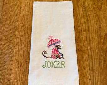 May include: White hand towel with an embroidered monkey wearing a pink and red outfit and holding a pink umbrella. The word "JOKER" is embroidered in green below the monkey.