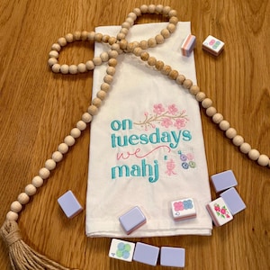 May include: A white linen towel embroidered with the phrase "on Tuesdays we mahj" in teal script, with pink floral accents. Several mahjong tiles are scattered around the towel, along with a wooden bead garland.