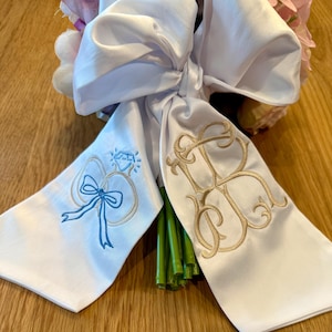 May include: White satin ribbon tied around a bouquet of flowers. One ribbon end features a blue bow and a diamond ring design, the other has gold embroidered initials. The bouquet has pink and purple flowers.