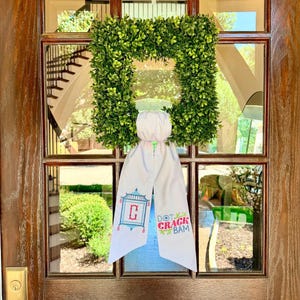 May include: A square green boxwood wreath featuring a white fabric ribbon hanging from the center. The ribbon displays a blue pagoda graphic with a red letter C and the text DOT CRACK BAM in pink and blue lettering with small green leaf accents.