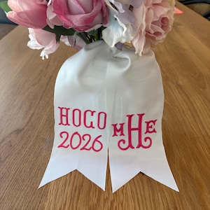 May include: A white ribbon hair accessory featuring pink embroidered text that reads HOCO 2026 on the left tail and a monogram with the letters MHE on the right tail, positioned beneath a bouquet of pink, white, and purple artificial flowers.