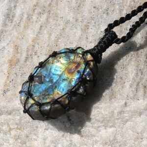 Raw Labradorite pendant displaying vivid flashes of electric blue, turquoise, gold, and hints of green across its natural rough surface. The stone is wrapped in a black macrame net with an adjustable black cord with a sliding knot closure and crystal beads and displayed on a light stone background. Labradorite is associated with transformation, intuition, protection, inner strength, spiritual awakening,  and enhanced creativity.