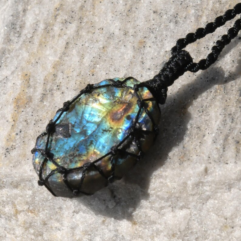 Raw Labradorite pendant displaying vivid flashes of electric blue, turquoise, gold, and hints of green across its natural rough surface. The stone is wrapped in a black macrame net with an adjustable black cord with a sliding knot closure and crystal beads and displayed on a light stone background. Labradorite is associated with transformation, intuition, protection, inner strength, spiritual awakening,  and enhanced creativity.