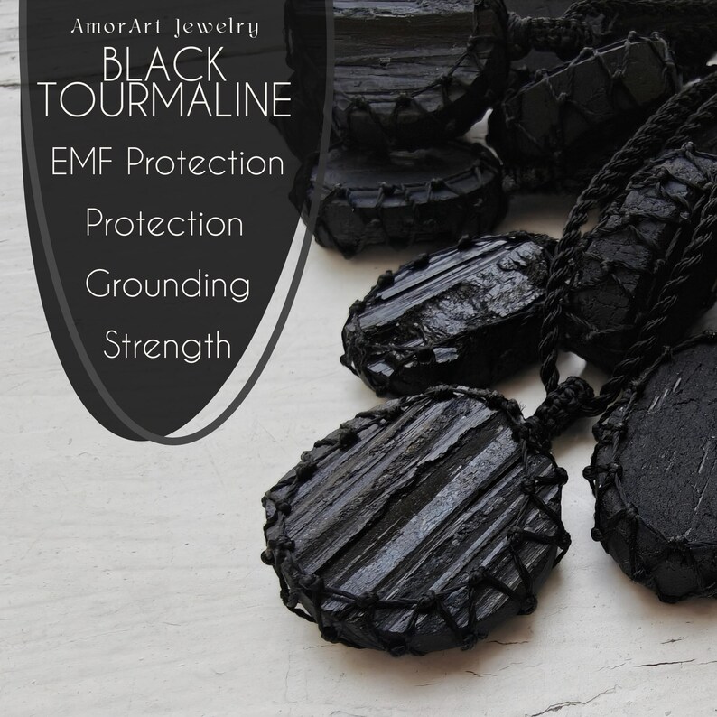 Close-up of multiple raw oval and round Black Tourmaline in deep black shades, resting on a white wood surface. The Black Tourmaline dark gemstone has strong parallel vertical striations, rough uneven edges, and a matte to slightly reflective texture with natural ridges and layered surfaces. Each piece is wrapped in a black macrame net and attached to a black cord with an adjustable knot closure. The image includes the text “Black Tourmaline, EMF Protection, Protection, Grounding, Strength”.