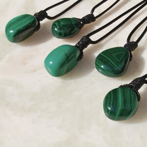 Close-up of 5 polished irregular Malachite in shades of vivid green and deep forest green, arranged on a light stone surface. The green opaque gemstones have smooth glossy surfaces with distinct banded patterns, concentric lines, and varied formations, including rounded and triangular shapes with layered green stripes and darker swirls. It is attached to a black cord with a top knot and adjustable knot closure. Malachite is associated with transformation, protection, and emotional healing