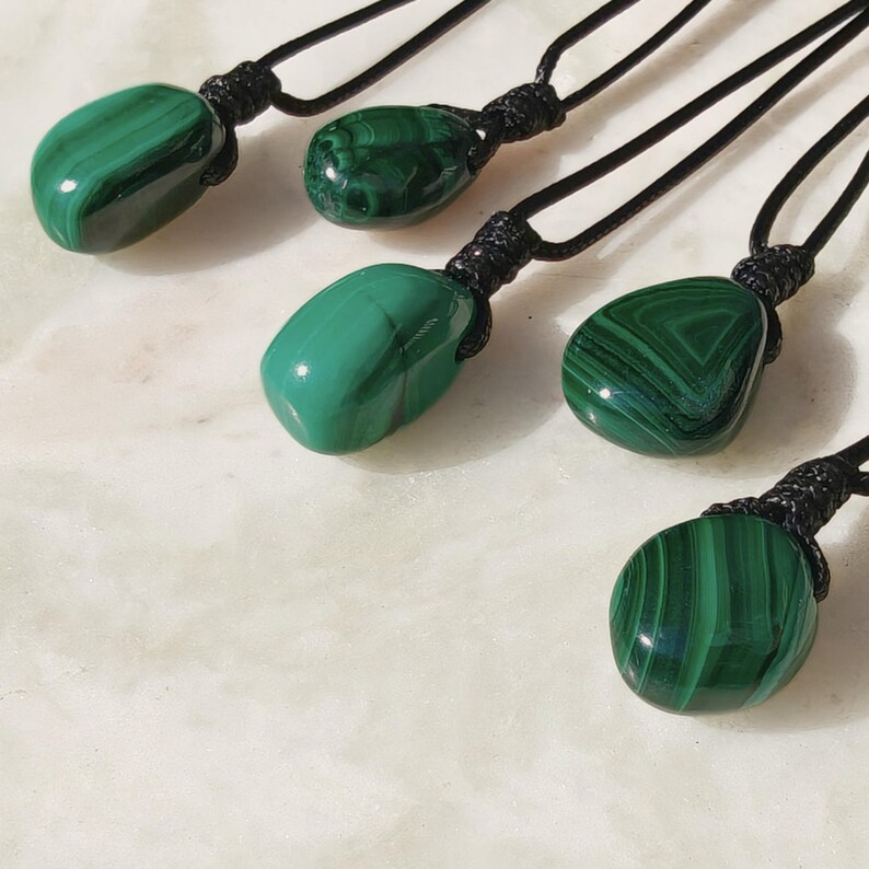 Close-up of 5 polished irregular Malachite in shades of vivid green and deep forest green, arranged on a light stone surface. The green opaque gemstones have smooth glossy surfaces with distinct banded patterns, concentric lines, and varied formations, including rounded and triangular shapes with layered green stripes and darker swirls. It is attached to a black cord with a top knot and adjustable knot closure. Malachite is associated with transformation, protection, and emotional healing