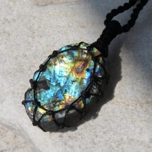 Raw Labradorite pendant displaying vivid flashes of electric blue, turquoise, gold, and hints of green across its natural rough surface. The stone is wrapped in a black macrame net with an adjustable black cord with a sliding knot closure and crystal beads and displayed on a light stone background. Labradorite is associated with transformation, intuition, protection, inner strength, spiritual awakening,  and enhanced creativity.