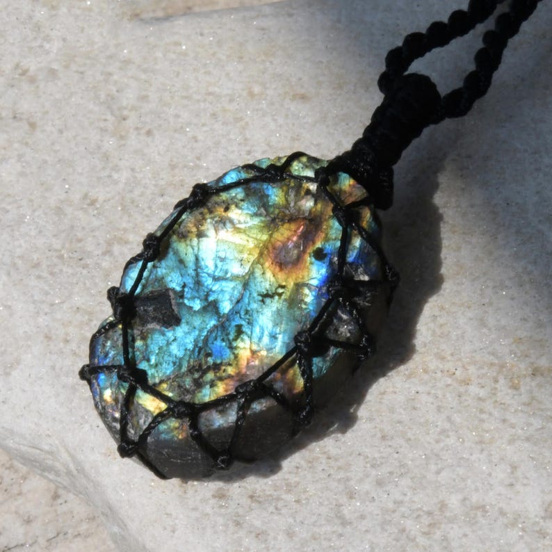 Raw Labradorite pendant displaying vivid flashes of electric blue, turquoise, gold, and hints of green across its natural rough surface. The stone is wrapped in a black macrame net with an adjustable black cord with a sliding knot closure and crystal beads and displayed on a light stone background. Labradorite is associated with transformation, intuition, protection, inner strength, spiritual awakening,  and enhanced creativity.