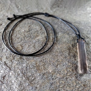 One raw smoky quartz necklace with rectangular stone in dark brown translucent hues, rough-textured with irregular edges, hung on a knotted black cord, placed on a light brown stone surface with a natural grainy texture. Smokey Quartz is associated with grounding, protection, and emotional calmness.