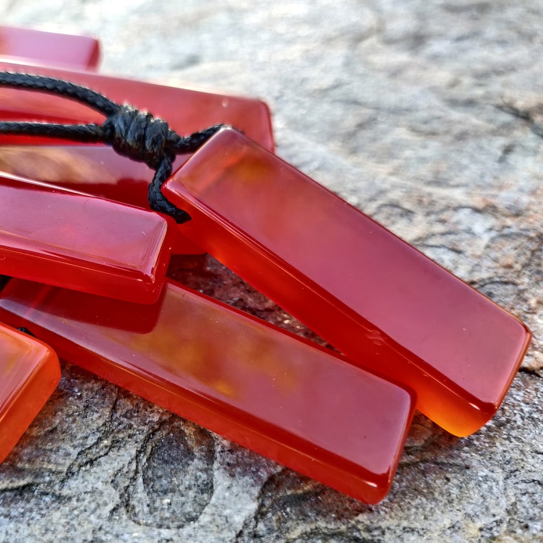 Several polished, elongated, rectangular Carnelian crystal necklaces are displayed on a textured gray surface. The orange-red gemstones have a smooth, translucent texture. There are multiple pendants, each attached to a black cord. Carnelian is associated with courage, vitality, and motivation.