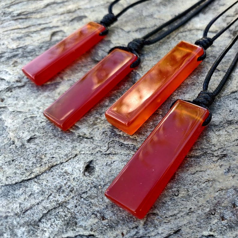 Four polished, elongated, rectangular Carnelian crystal necklaces are displayed on a textured gray surface. The orange-red gemstones have a smooth, translucent texture. There are multiple pendants, each attached to a black cord. Carnelian is associated with courage, vitality, and motivation.