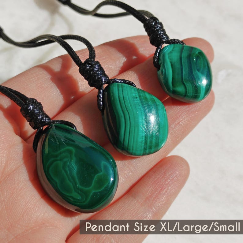 Close-up of 3 polished irregular Malachite in shades of vivid green and deep forest green, displayed on a hand against a light background. The green opaque gemstones have smooth glossy surfaces with distinct banded patterns, flowing lines, and concentric formations, with light reflections highlighting their curved shapes and surface polish. It is attached to a black cord with a top knot and adjustable knot closure. The image includes the text Pendant Size XL Large Small.