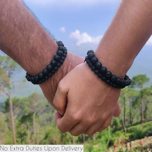 Pode incluir: Duas pulseiras de contas de pedra de lava preta são usadas nos pulsos, com as mãos unidas. As pulseiras são feitas de pequenas contas redondas e escuras. O fundo mostra uma paisagem desfocada com árvores e céu azul.