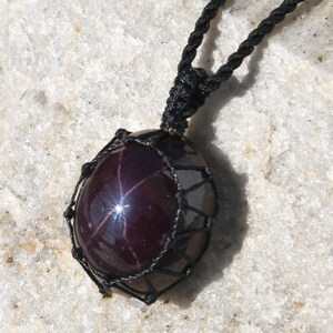 Close-up of a polished star garnet pendant in deep burgundy, wine-red, and dark plum tones, displayed on a light stone background. A distinct four-rayed star appears across the gemstone when illuminated, creating a bright reflective cross. The garnet has a smooth glossy finish with rich color depth and natural translucency. It is secured in a black macrame net and attached to a black adjustable cord. Associated with passion, vitality, grounding, courage, and emotional strength.